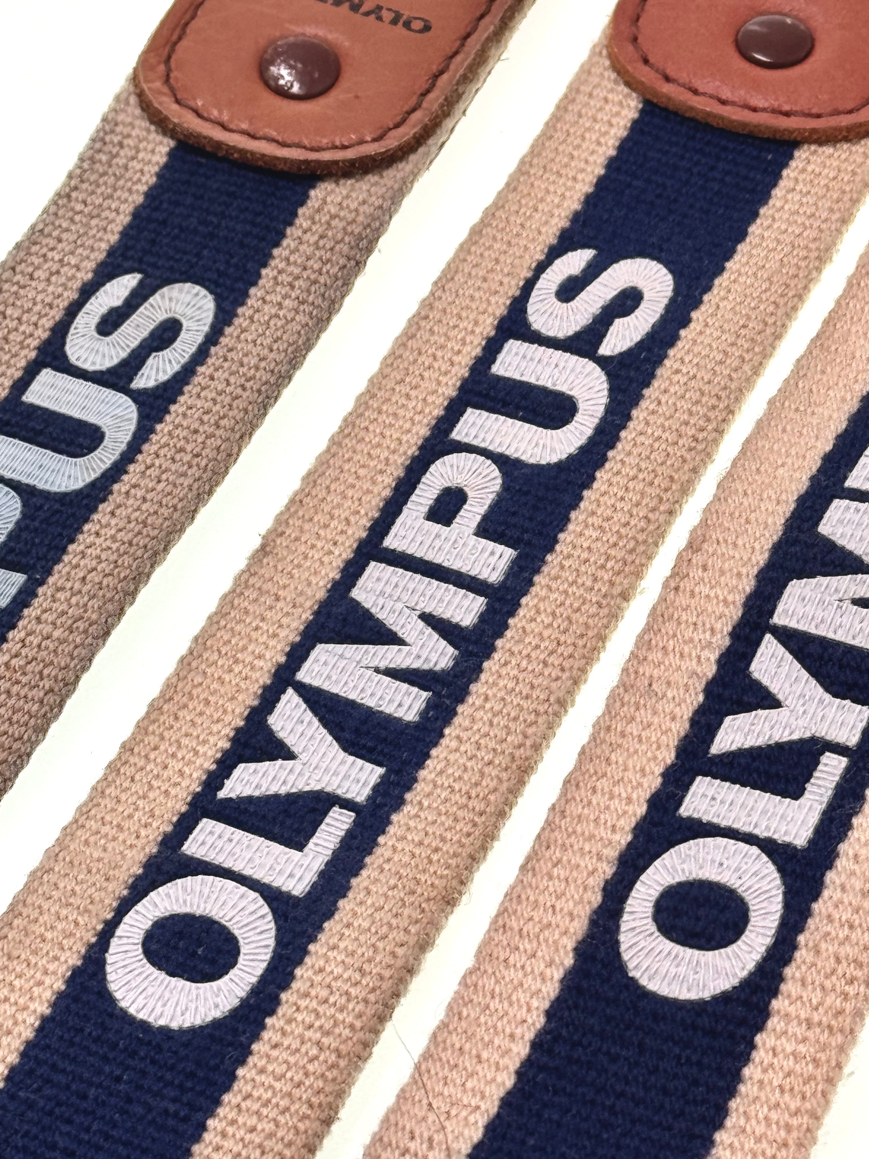 Close-up of a striped camera strap with 'Olympus' branding on a white background - The Olympus Hunter Strap